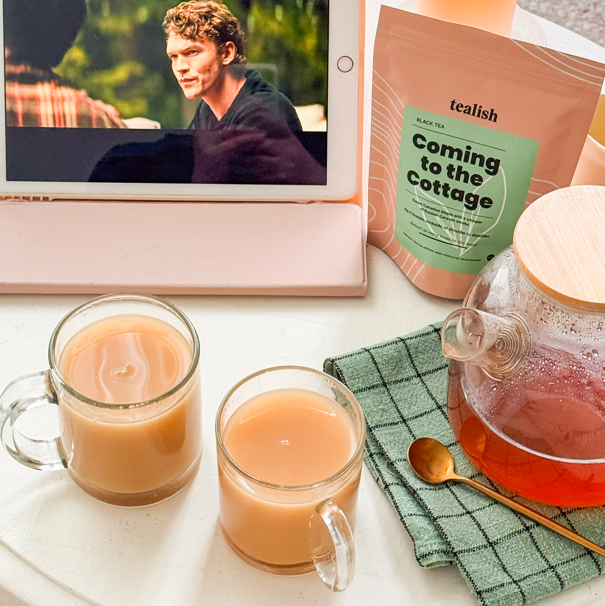 Table with a tablet showing a movie, two cups of tea, a teapot, and a bag of icalish tea.
