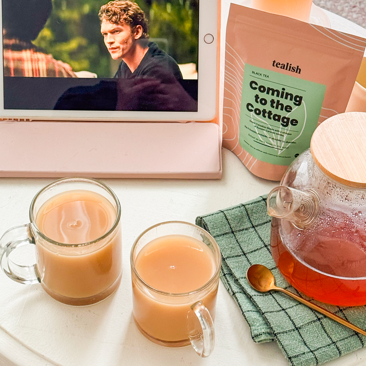 Table with a tablet showing a movie, two cups of tea, a teapot, and a bag of icalish tea.
