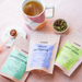 Tea bags labeled 'Sacred Pause', 'Moonbrew', and 'First Light' with a mug of tea on a pink surface.
