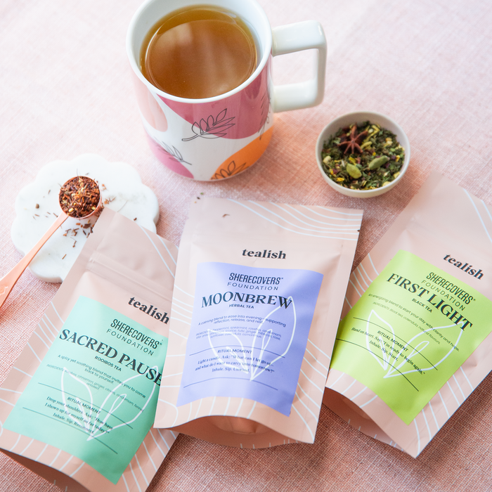 Tea bags labeled 'Sacred Pause', 'Moonbrew', and 'First Light' with a mug of tea on a pink surface.