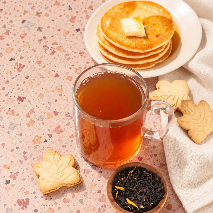 A clear glass mug filled with amber tea sits on a speckled pink surface, with a plate of golden pancakes topped with butte and maple syrupr, maple leaf-shaped cookies, and a small bowl of loose leaf tea nearby.