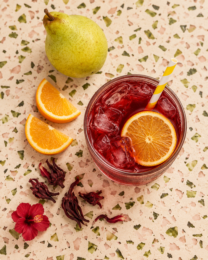 A glass of iced red hibiscus tea with orange slices and a yellow-and-white striped straw, arranged on a speckled beige surface with a pear, orange wedges, dried hibiscus flowers, and a red flower in the frame.