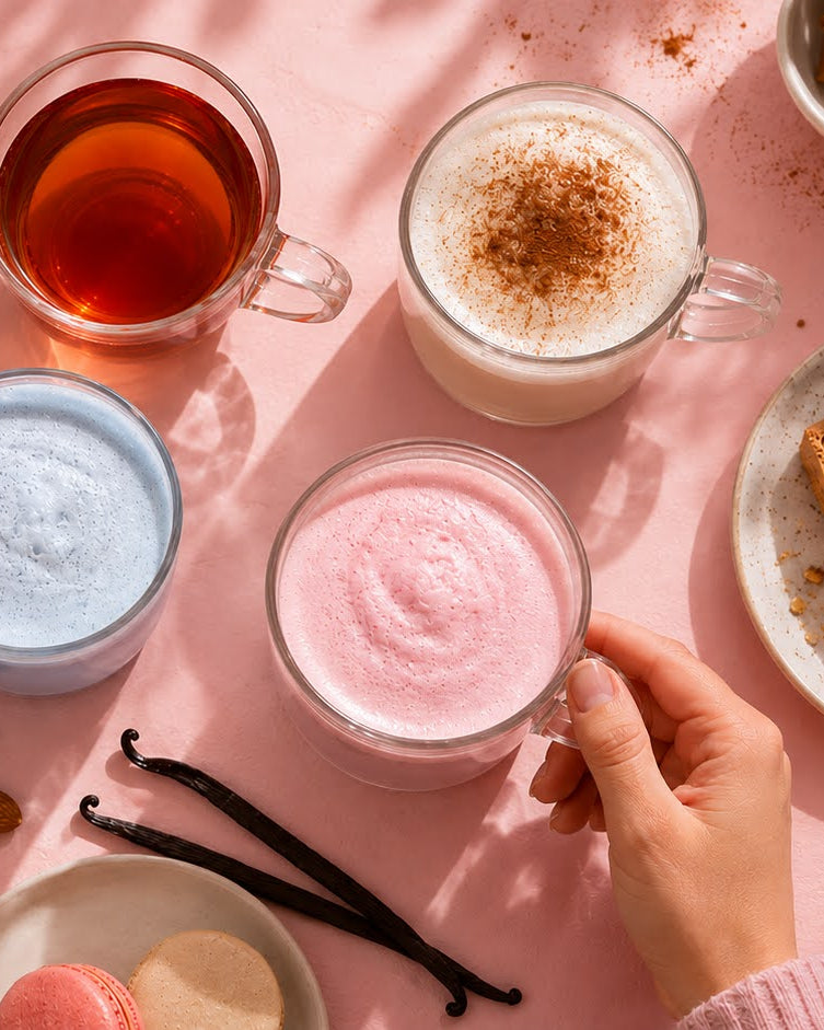 A top-down styled flat lay of assorted beverages and desserts on a pink surface. Visible items include a slice of meringue pie, biscotti, cake pieces, macarons, cinnamon sticks, and four drinks in glass cups and mugs: an amber tea, a light blue frothy drink, a pink frothy drink being held by a hand, and a white drink with cocoa powder on top.