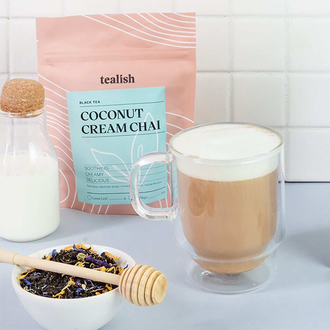 A package of Tealish Coconut Cream Chai tea next to a glass mug filled with tea latte and milk, with a bowl of spices and a honey dipper in front, all set against a white tiled background.