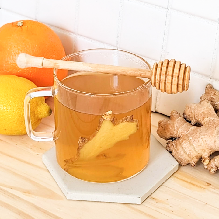 A clear glass mug filled with ginger tea, alongside a honey dipper and lemon, with ginger roots visible in the background.