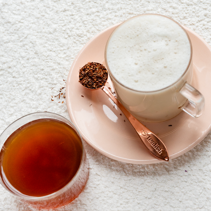 A cup of rooibos tea with a swirl of cream on top, alongside a small bowl of rooibos leaves and a spoon, on a pink plate on a white surface.