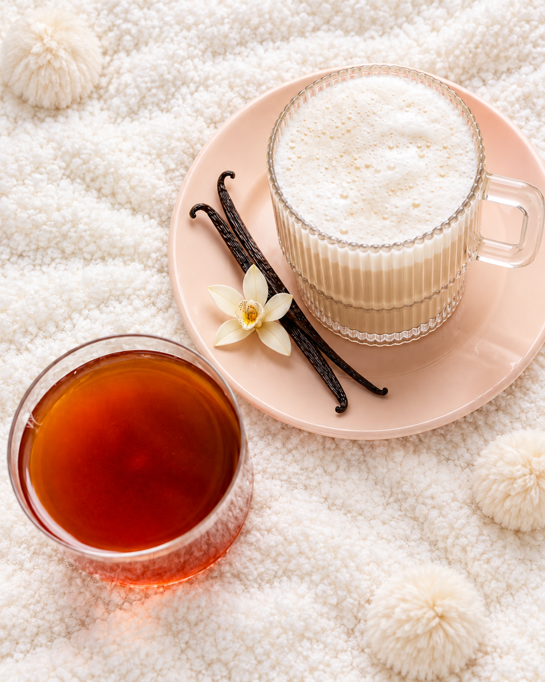 A styled flat-lay of two clear glass cups on a soft white textured blanket. One cup holds a frothy light beige beverage on a pale pink saucer with vanilla beans and a small white flower.