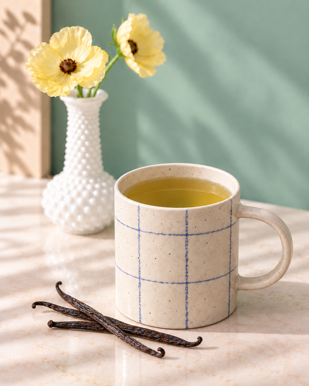 A speckled cream ceramic mug with a blue grid pattern, filled with vanilla green tea, sits on a marble surface next to vanilla beans, with a white textured vase of yellow flowers in the background.