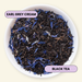 A top-down view of loose-leaf tea in a white bowl, featuring dark black tea leaves mixed with blue flower petals. Two lavender labels with black text are overlaid: 'EARL GREY CREAM' near the top left and 'BLACK TEA' near the bottom right, set against a pale cream background.