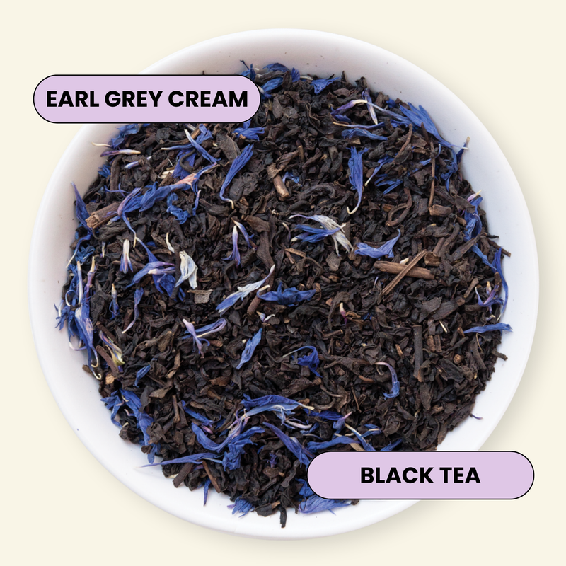 A top-down view of loose-leaf tea in a white bowl, featuring dark black tea leaves mixed with blue flower petals. Two lavender labels with black text are overlaid: 'EARL GREY CREAM' near the top left and 'BLACK TEA' near the bottom right, set against a pale cream background.