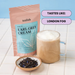 A stand-up pouch of tealish Earl Grey Cream black tea stands next to a frothy latte in a glass mug and a small bowl of loose tea leaves on a tiled surface, with text bubbles reading 'TASTES LIKE: LONDON FOG'.