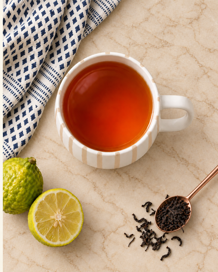 A white ceramic mug filled with amber tea sits on a beige marble surface, styled with a blue-and-white checkered cloth, a whole lime, a lime half, loose tea leaves, and a copper spoon holding tea leaves.