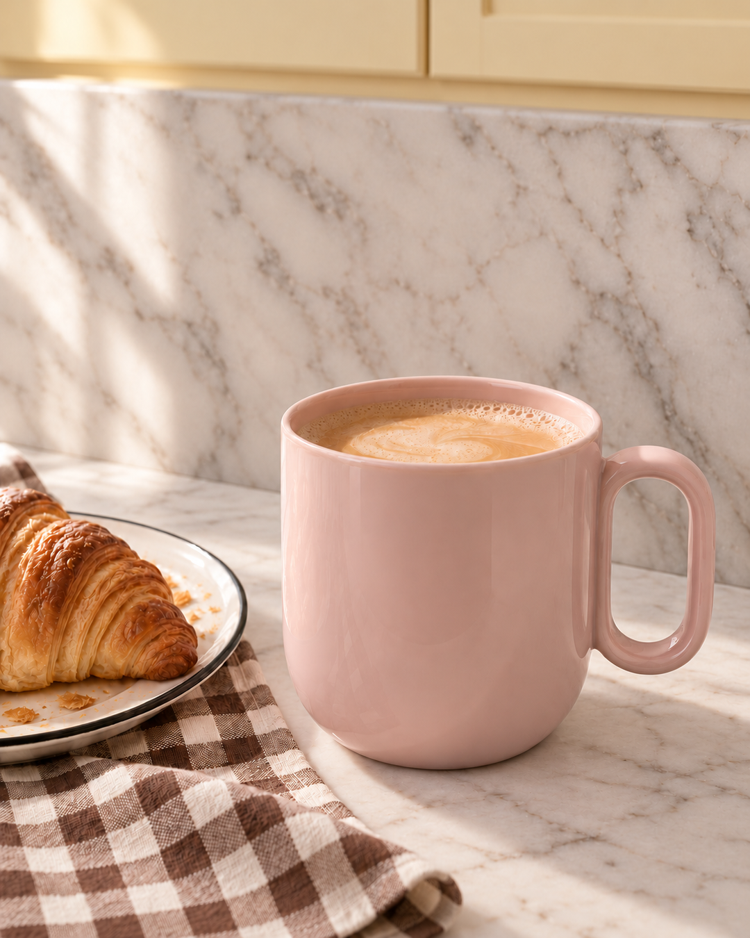 A dusty pink ceramic tea mug filled with a tea latte, set on a marble countertop next to a croissant on a checkered napkin.