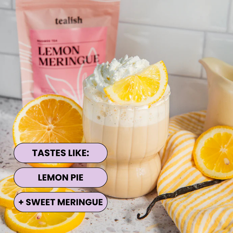 A glass of frothy lemon meringue tea topped with whipped cream and a lemon slice, with sliced lemons, a striped cloth, and a pink tea pouch labeled tealish Lemon Meringue in the background. Text on the image says it tastes like lemon pie and sweet meringue.