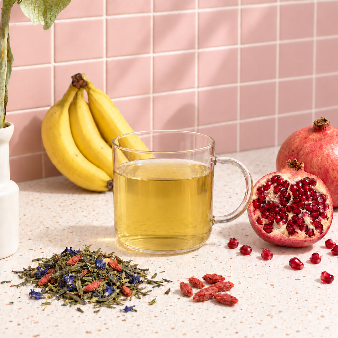 A clear glass mug filled with green tea sits on a speckled countertop, surrounded by loose herbal tea, red goji berries, a halved pomegranate, and a bunch of bananas against a pink tiled wall.