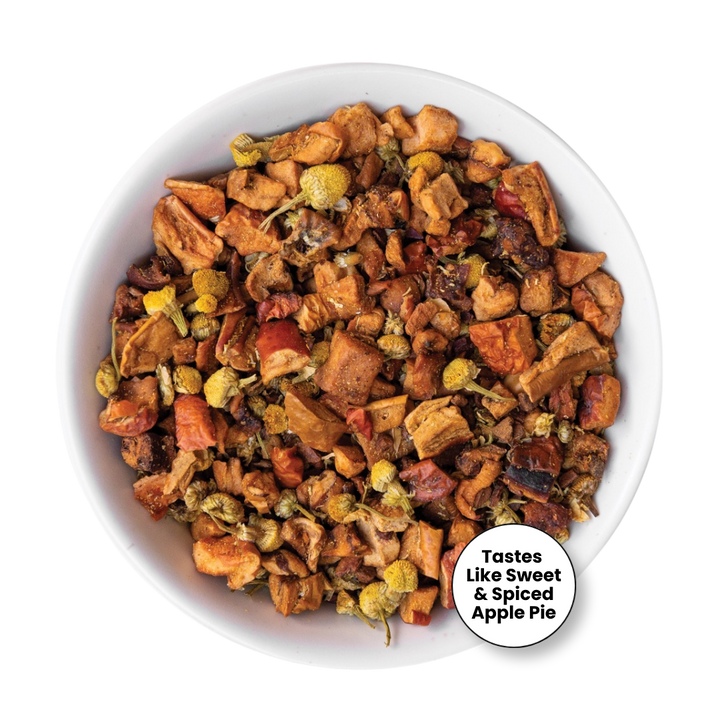 A top-down view of a white bowl filled with a loose-leaf herbal tea blend. The tea contains pieces of dried apple, orange peel, and small yellow flower buds.