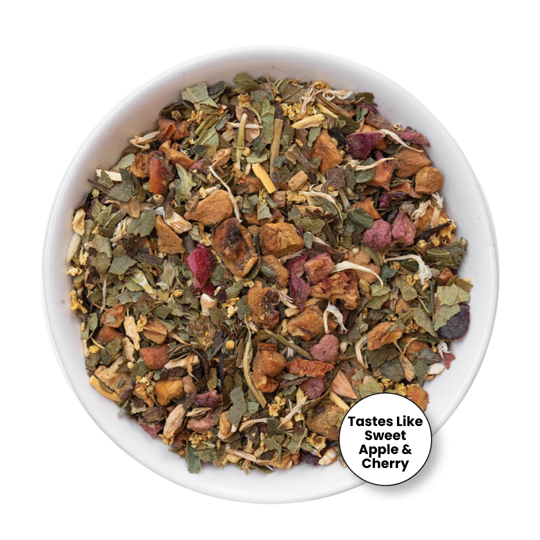 A top-down view of a white bowl filled with loose-leaf herbal tea blend, featuring dried leaves, apple pieces, and cherry pieces, with a circular badge in the lower right that reads &
