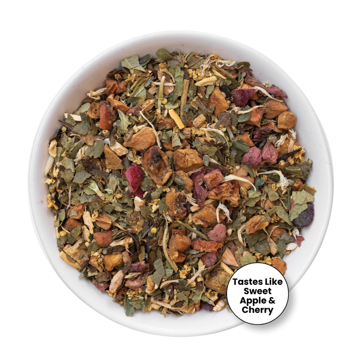 A top-down view of a white bowl filled with loose-leaf herbal tea blend, featuring dried leaves, apple pieces, and cherry pieces, with a circular badge in the lower right that reads &
