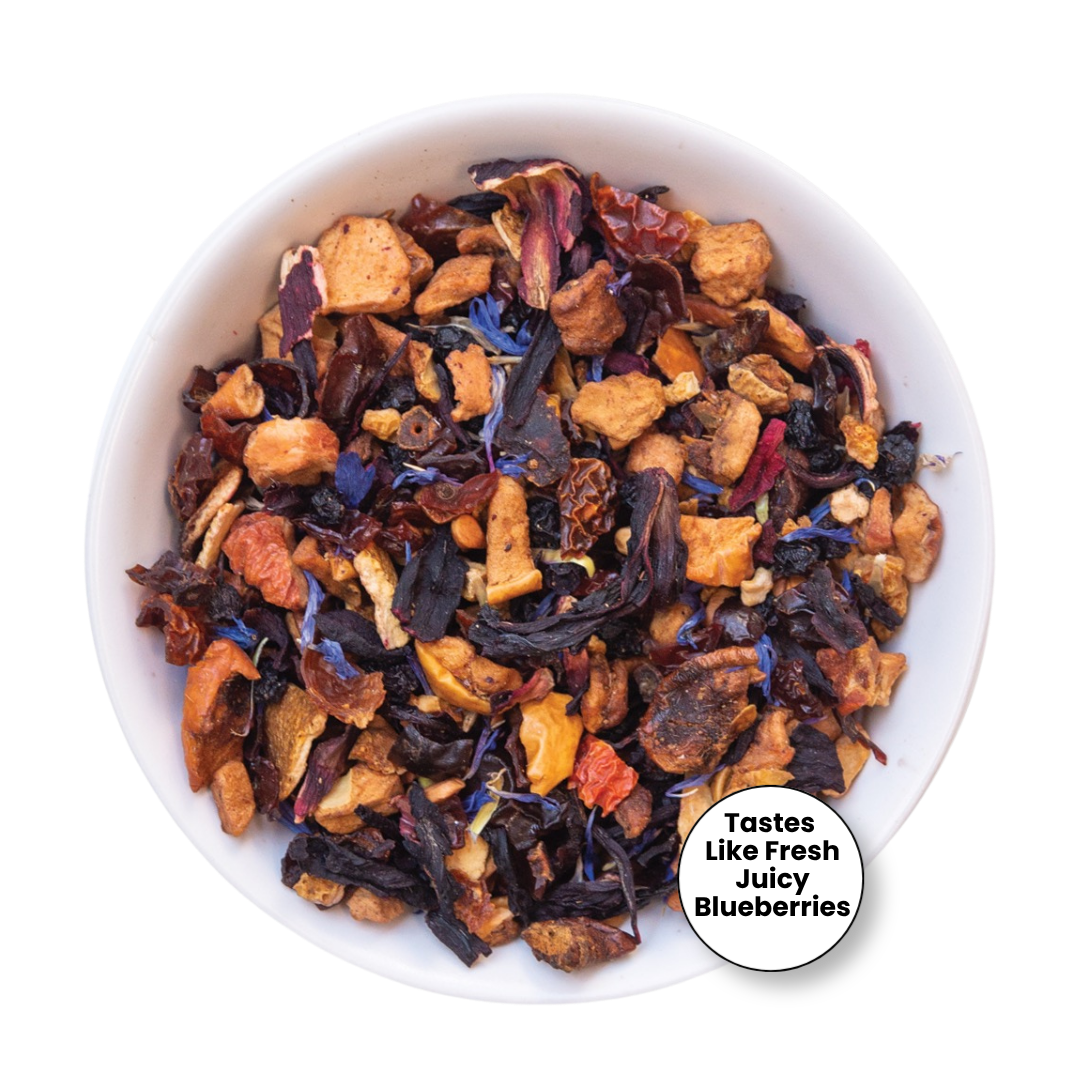 A top-down view of a white bowl filled with a loose-leaf herbal tea blend. The tea contains dried blueberries, orange citrus pieces, dark dried flowers, and small purple wildflower elements.