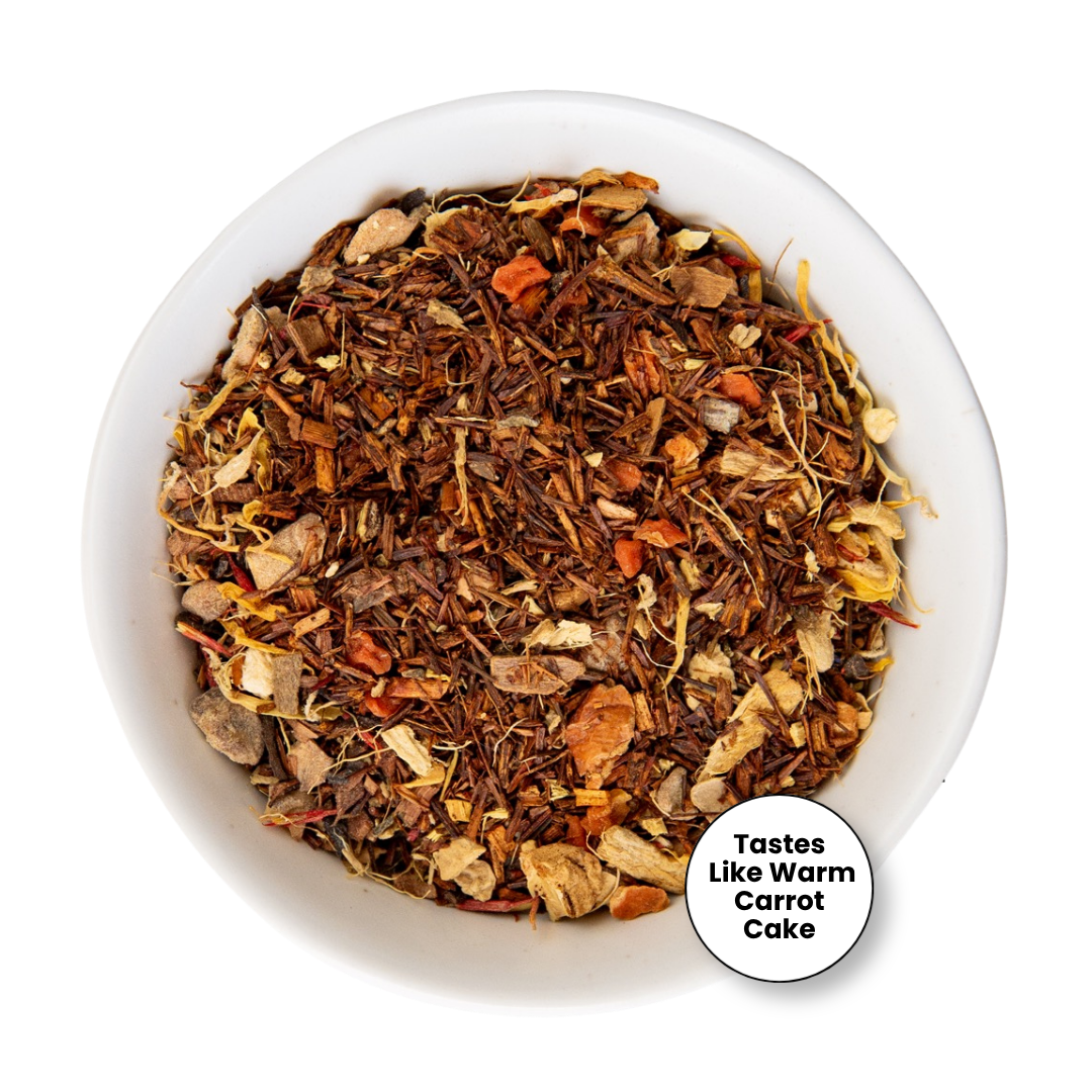 A top-down view of a white bowl filled with a loose-leaf herbal tea blend. The tea contains dark brown tea leaves, dried orange carrot pieces, and light tan dried fruit or flower pieces.