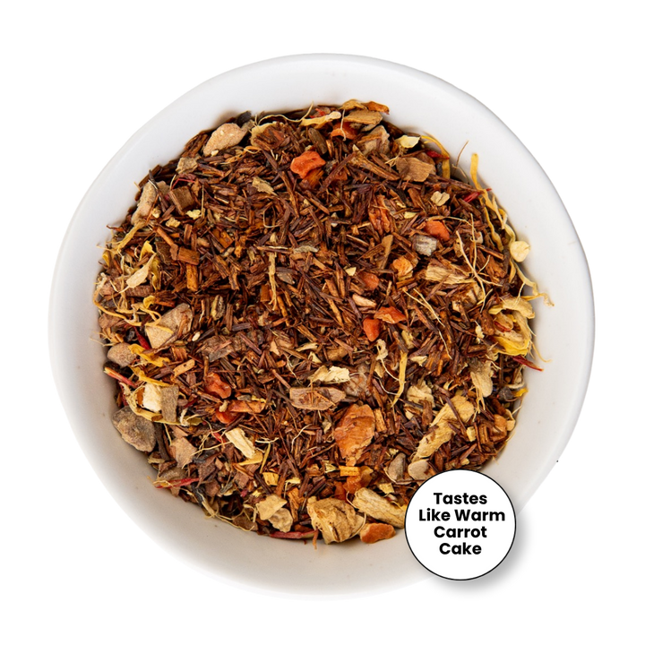 A top-down view of a white bowl filled with a loose-leaf herbal tea blend. The tea contains dark brown tea leaves, dried orange carrot pieces, and light tan dried fruit or flower pieces.