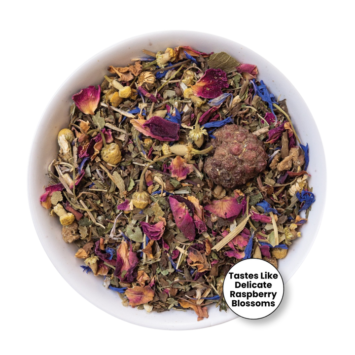 A white bowl filled with loose-leaf herbal tea blend featuring dried flowers, petals, herbs, and a round brown tea ball, with a white circular badge in the lower right reading &