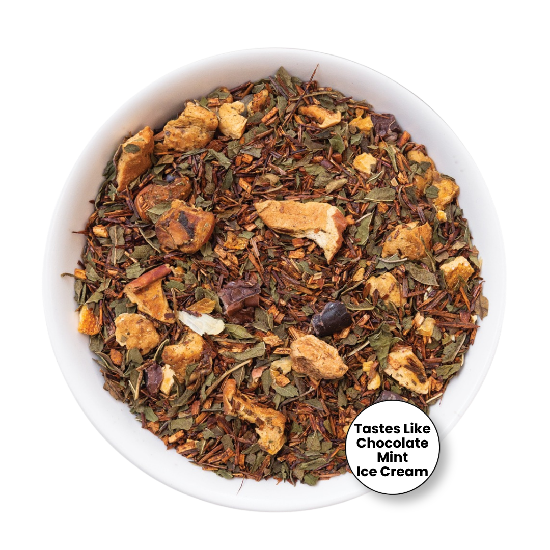A top-down view of a white bowl filled with a loose-leaf herbal tea blend. The tea contains dried green leaves, brown rosebuds, and tan dried fruit pieces.