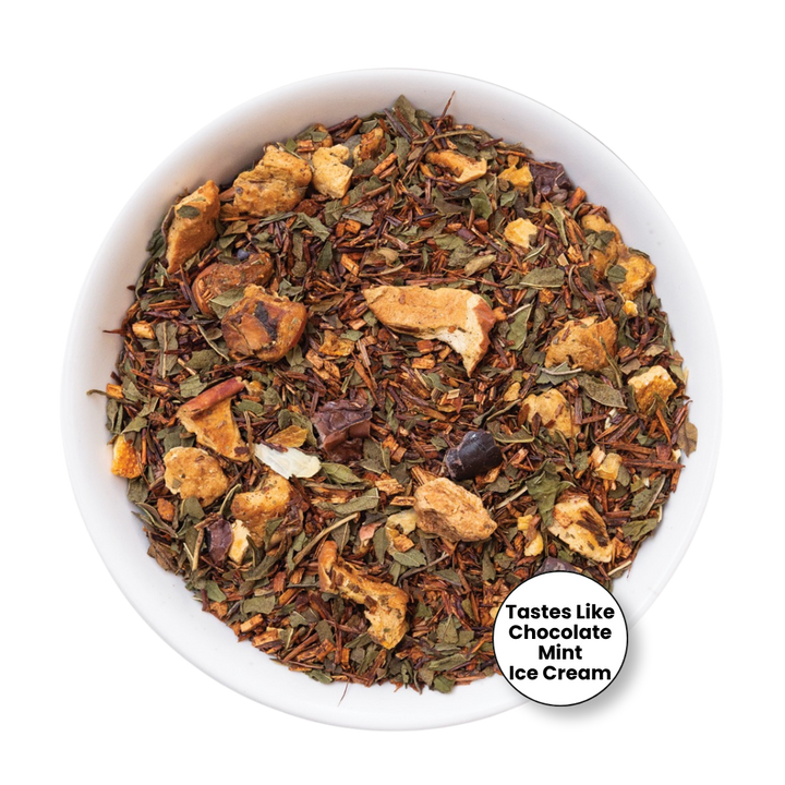A top-down view of a white bowl filled with a loose-leaf herbal tea blend. The tea contains dried green leaves, brown rosebuds, and tan dried fruit pieces.