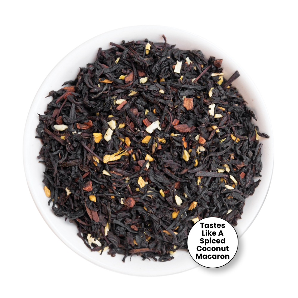 A top-down view of a loose-leaf tea blend in a white bowl, featuring dark dried tea leaves mixed with toasted coconut flakes and small dried fruit pieces. A white circular badge in the bottom right corner reads, 'Tastes Like A Spiced Coconut Macaron'.