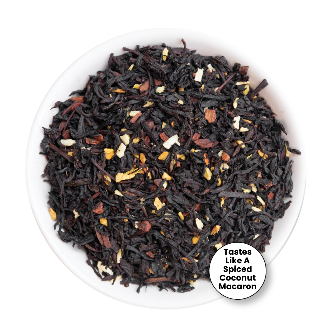 A top-down view of a loose-leaf tea blend in a white bowl, featuring dark dried tea leaves mixed with toasted coconut flakes and small dried fruit pieces. A white circular badge in the bottom right corner reads, &