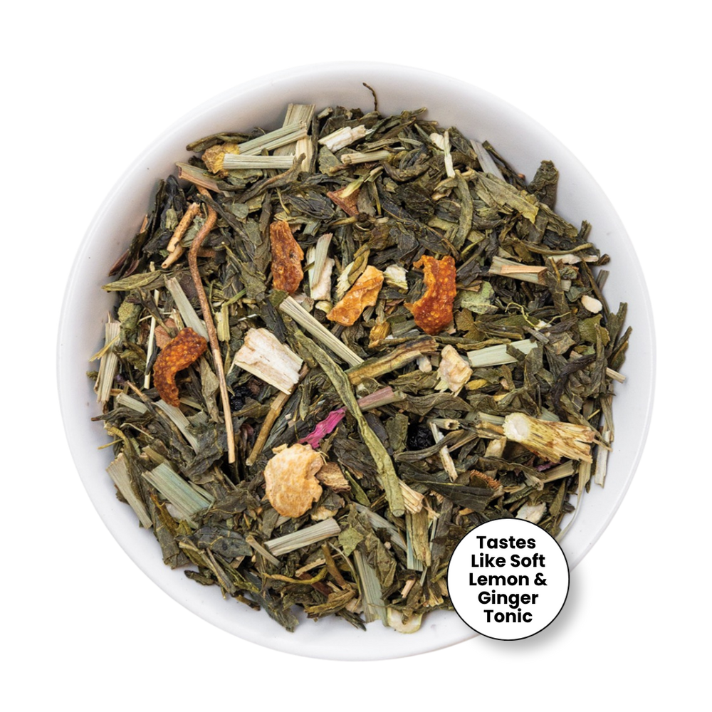 A top-down view of loose-leaf herbal tea inside a white bowl, featuring green leaves, dried orange pieces, and pale yellow flower petals, with a circular label that reads 'Tastes Like Soft Lemon & Ginger Tonic'.