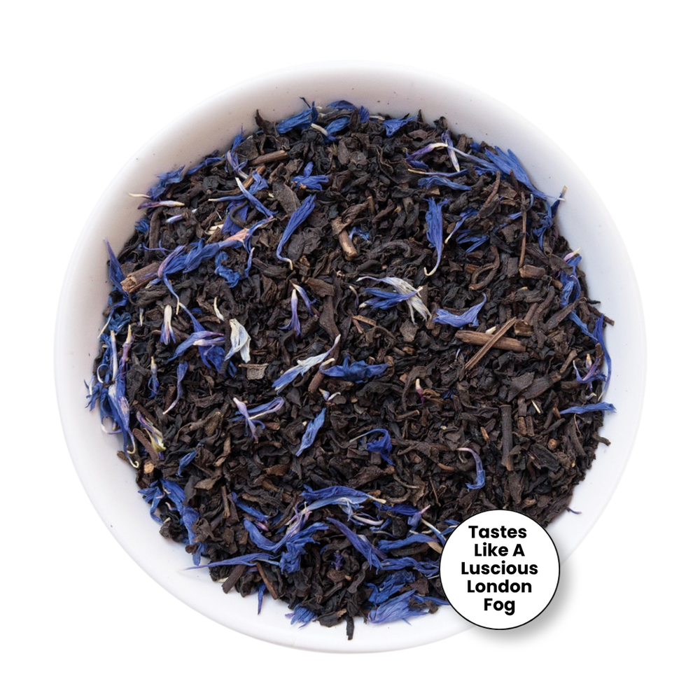 A top-down view of loose-leaf tea inside a white bowl, featuring dark brown tea leaves mixed with bright blue dried flower petals. A white circular badge in the bottom right corner reads 'Tastes Like A Luscious London Fog'.