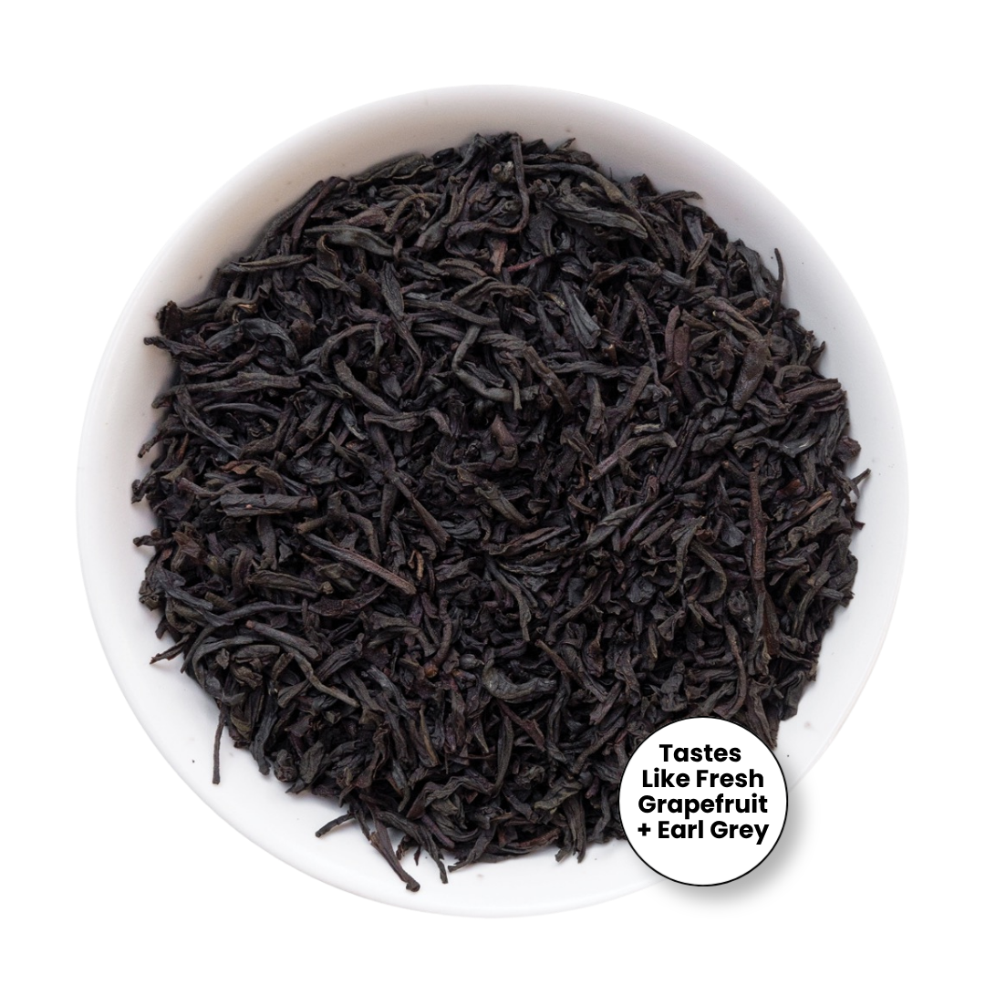 A top-down view of loose-leaf tea in a white bowl, with a small white circular label in the lower right that reads &