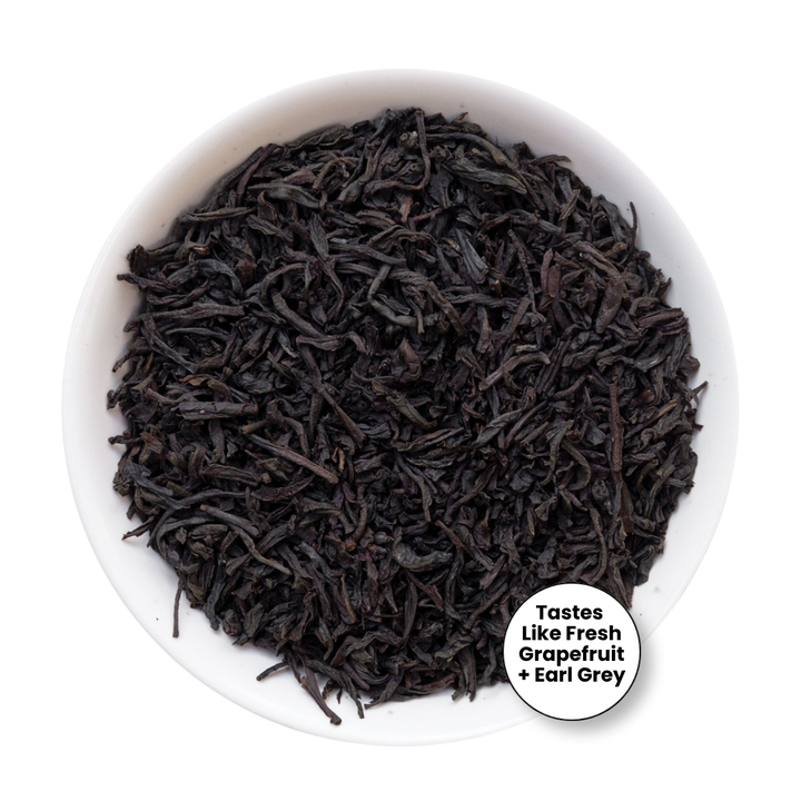 A top-down view of loose-leaf tea in a white bowl, with a small white circular label in the lower right that reads &