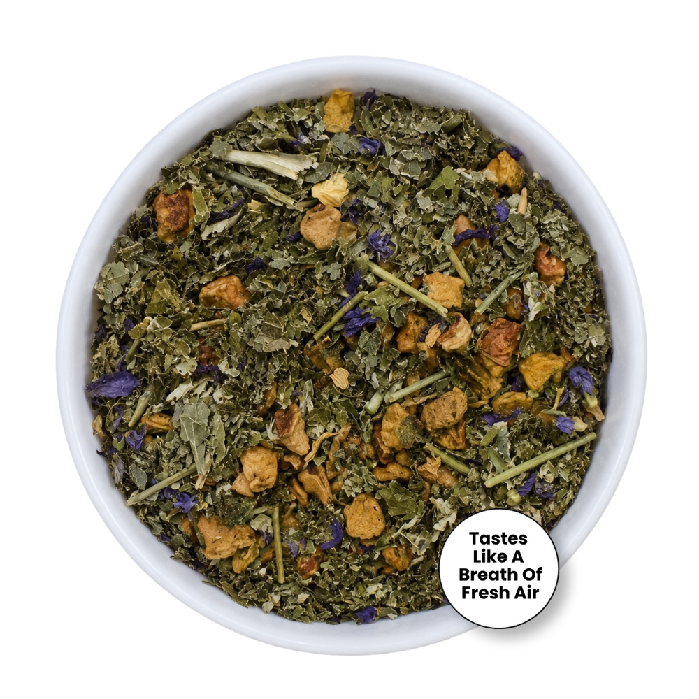 A top-down view of a white bowl filled with dried herbal tea blend, featuring green leaves, yellow-brown flower petals, and small purple accents, with a circular badge that reads 'Tastes Like A Breath Of Fresh Air'.