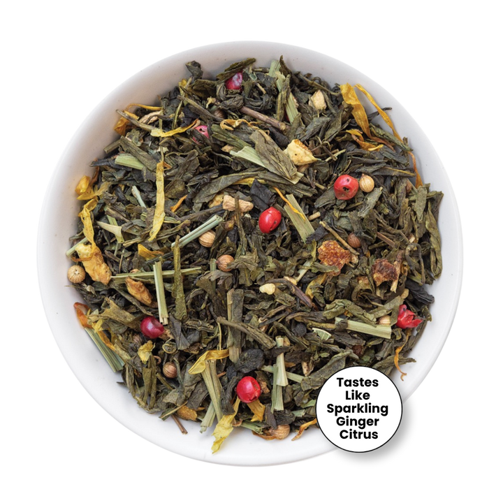A top-down view of a white bowl filled with loose-leaf herbal tea blend, featuring dried green leaves, yellow citrus peel, small brown pieces, and bright red berries. A circular badge in the lower right corner reads &