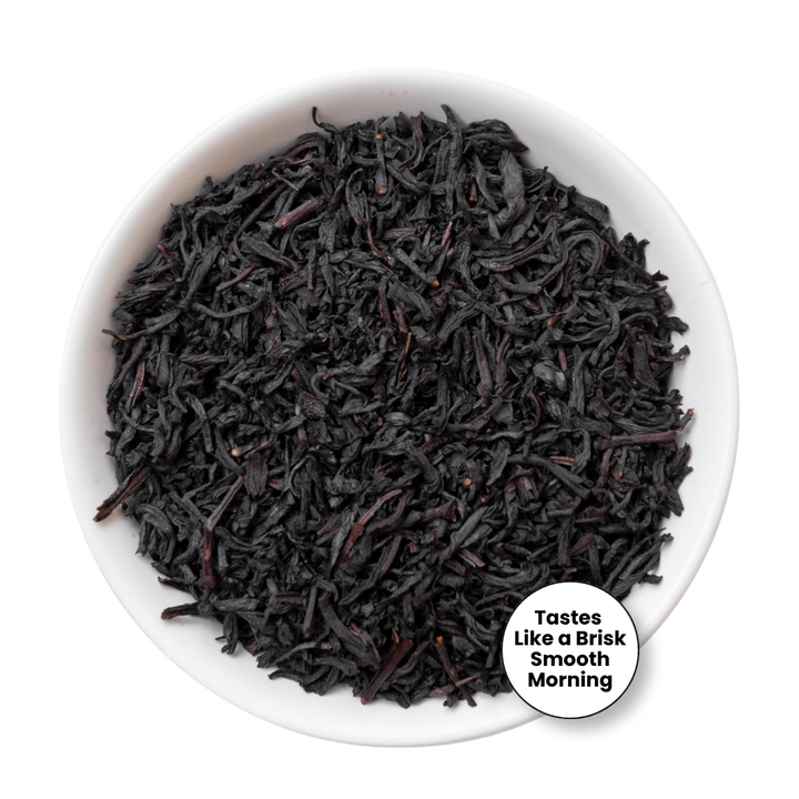 Dark black tea leaves piled in a white bowl, with a small circular badge in the lower right that reads &
