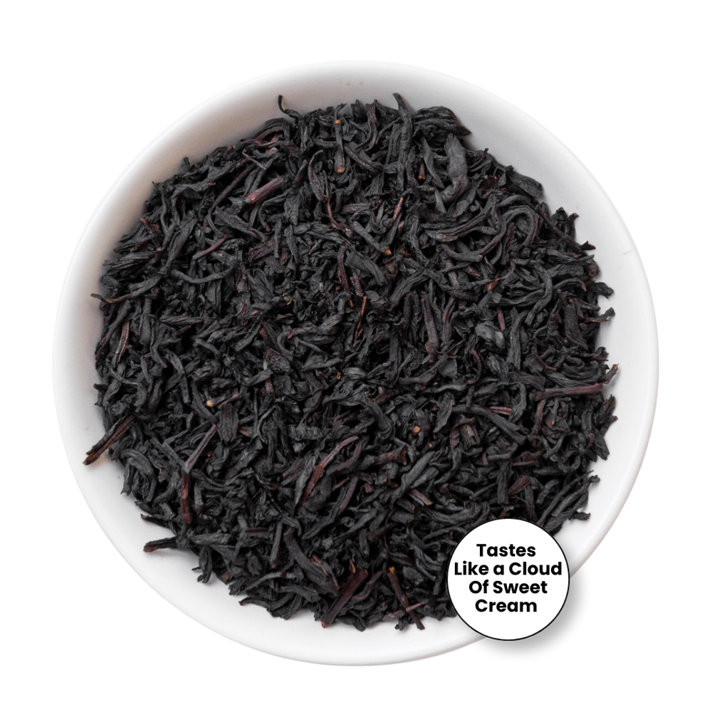 A top-down view of dark, curly tea leaves in a white bowl, with a white circular badge in the lower right that reads 'Tastes Like a Cloud Of Sweet Cream' on a plain white background.