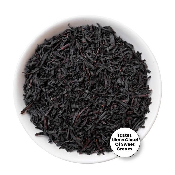 A top-down view of dark, curly tea leaves in a white bowl, with a white circular badge in the lower right that reads &
