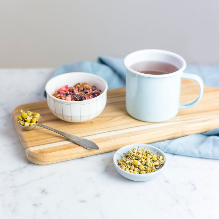 A cup of chamomile tea is placed on a wooden board with a bowl of granola next to it. There are also small bowls with what appears to be dried chamomile flowers.