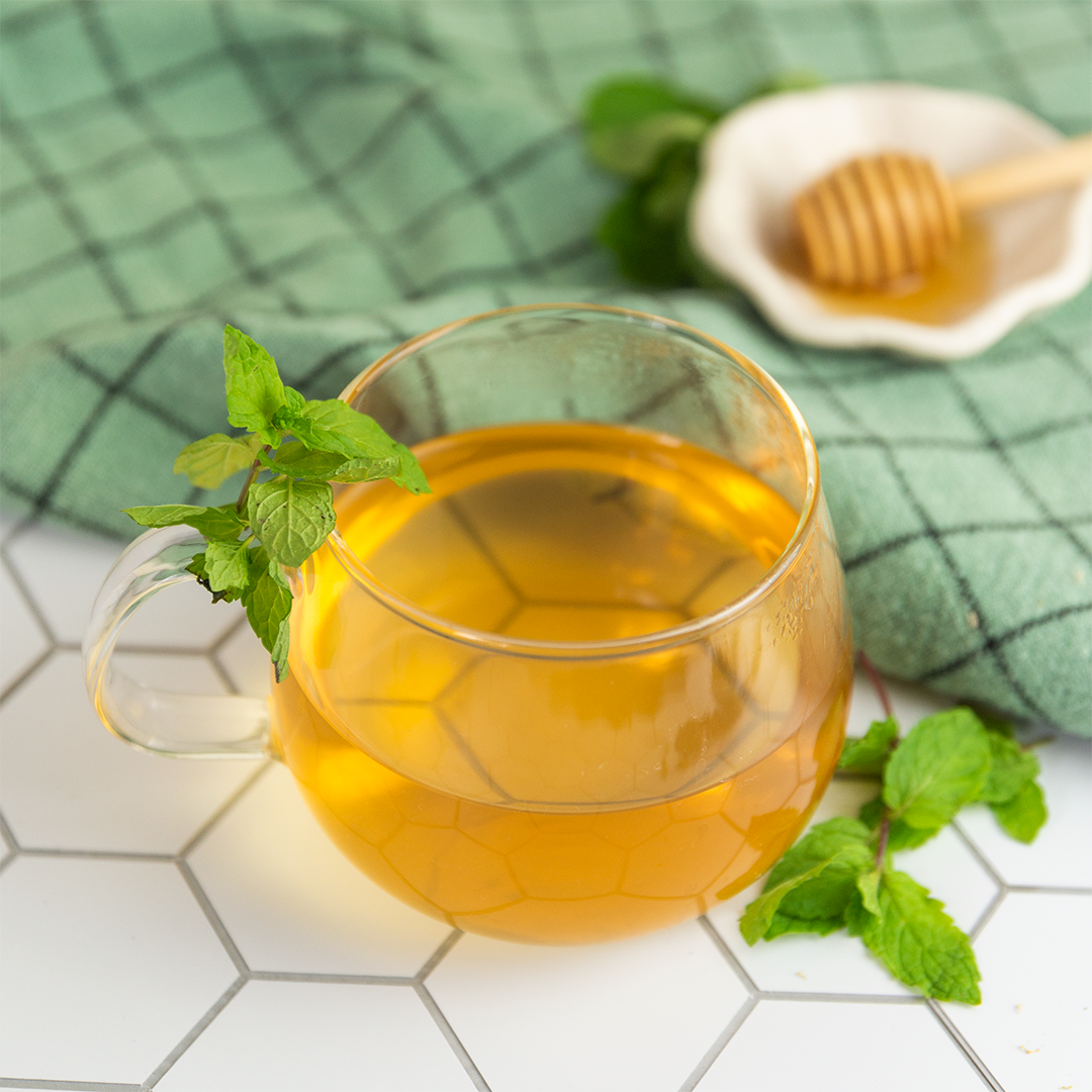A clear glass cup filled with peppermint tea, garnished with a sprig of mint, alongside a honey dish and fresh mint leaves on a green checkered napkin.
