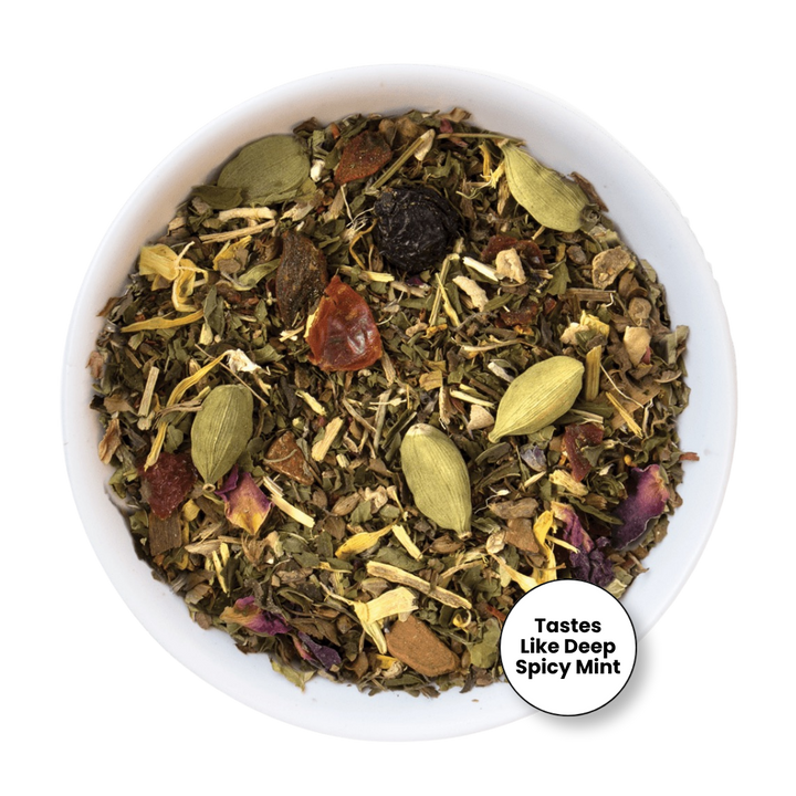 A top-down view of a white bowl filled with a loose herbal tea blend. The tea contains green cardamom pods, dried flowers, and small brown pieces.