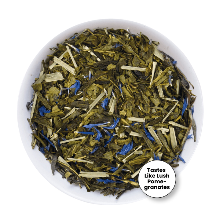 A top-down view of loose-leaf herbal tea in a white bowl, featuring dried green leaves, pale tan sticks, and small bright blue flower pieces. A circular badge in the lower right says &
