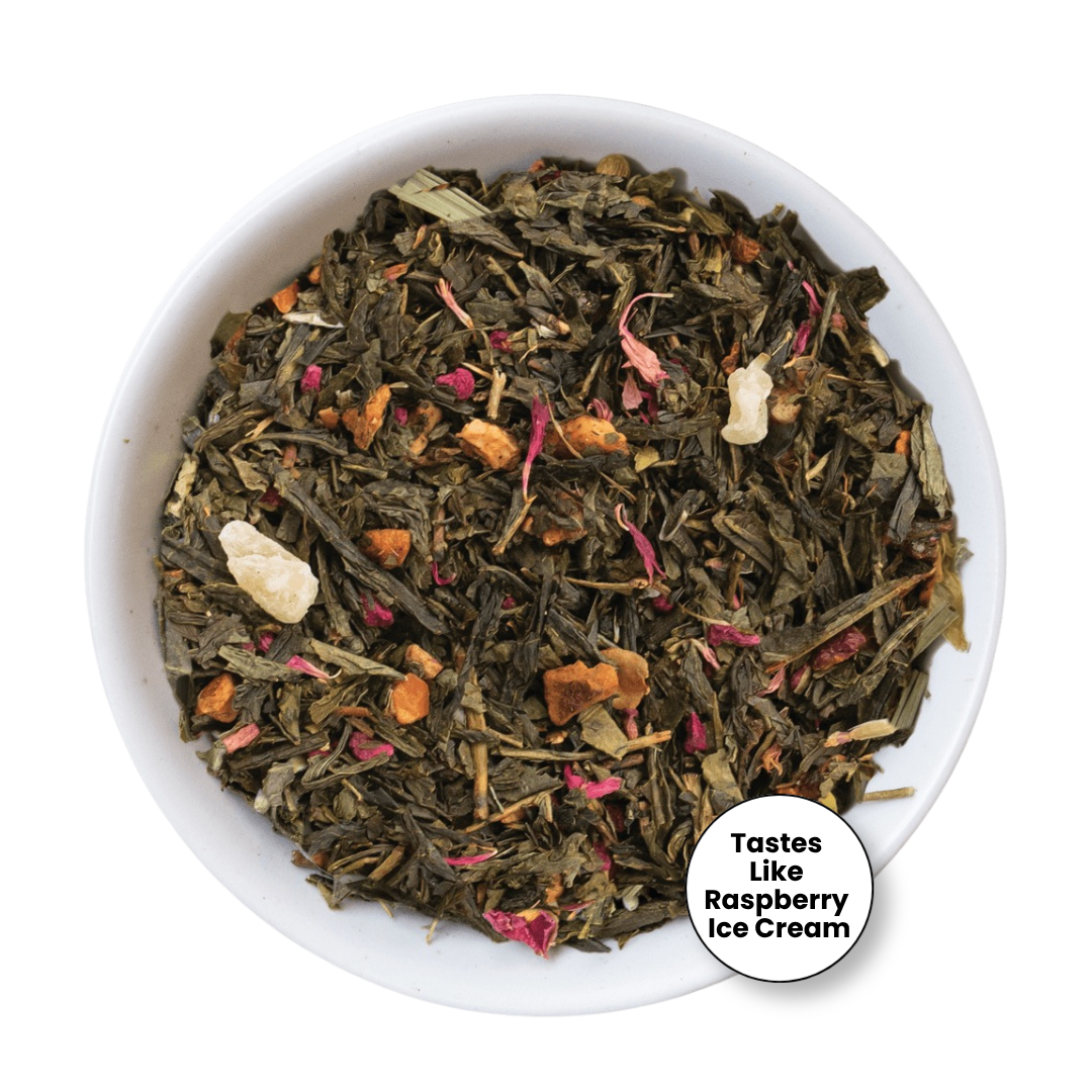 A top-down view of a white bowl filled with loose-leaf herbal tea blend. The tea contains dark green tea leaves, dried pink flower petals, orange dried fruit pieces, and a few pale beige chunks.