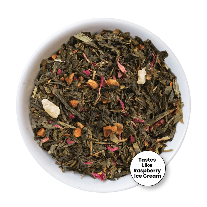 A top-down view of a white bowl filled with loose-leaf herbal tea blend. The tea contains dark green tea leaves, dried pink flower petals, orange dried fruit pieces, and a few pale beige chunks.