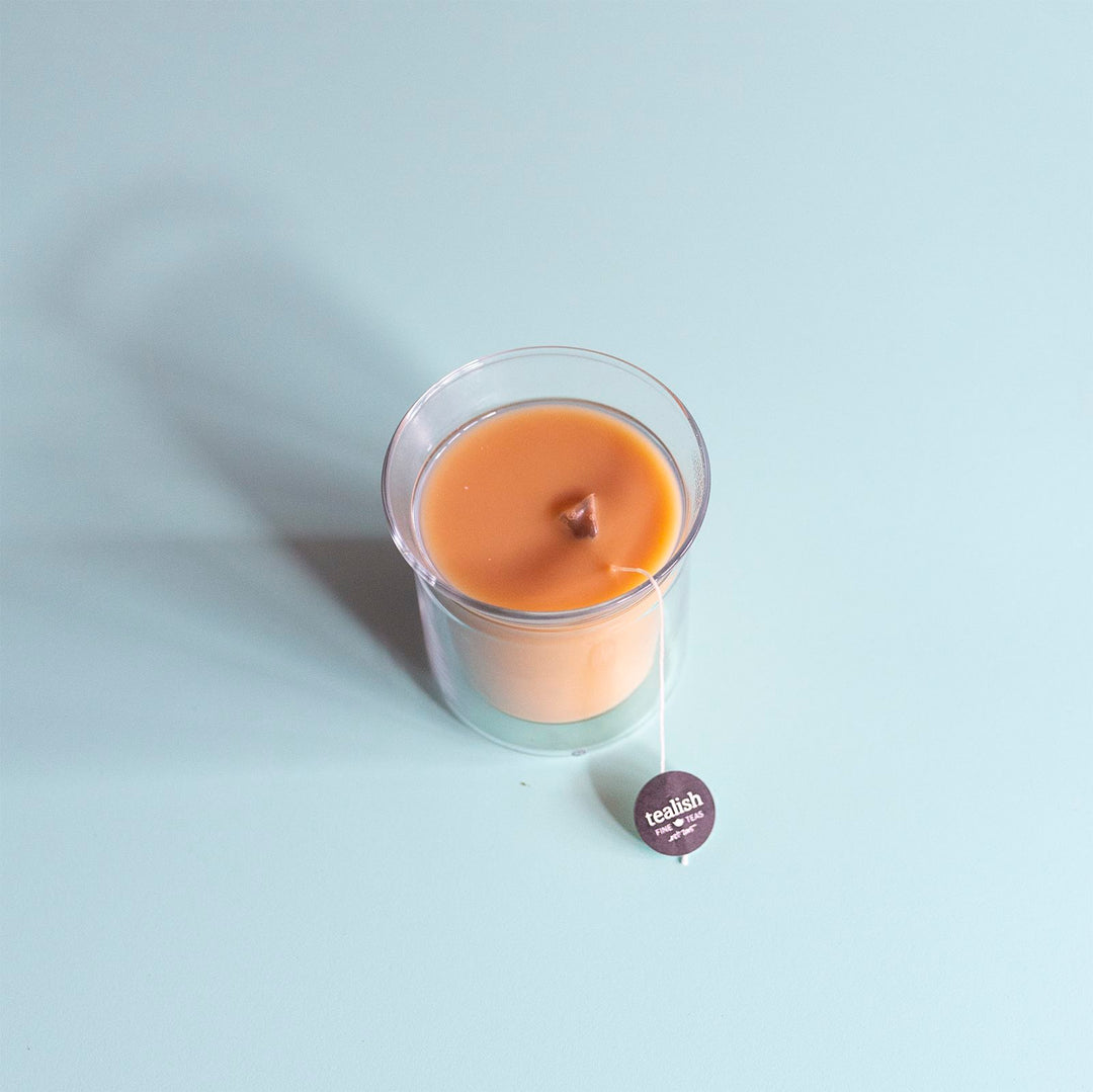A candle with a beige hue, possibly orange or light brown, in a clear glass container, with a tea bag marked &