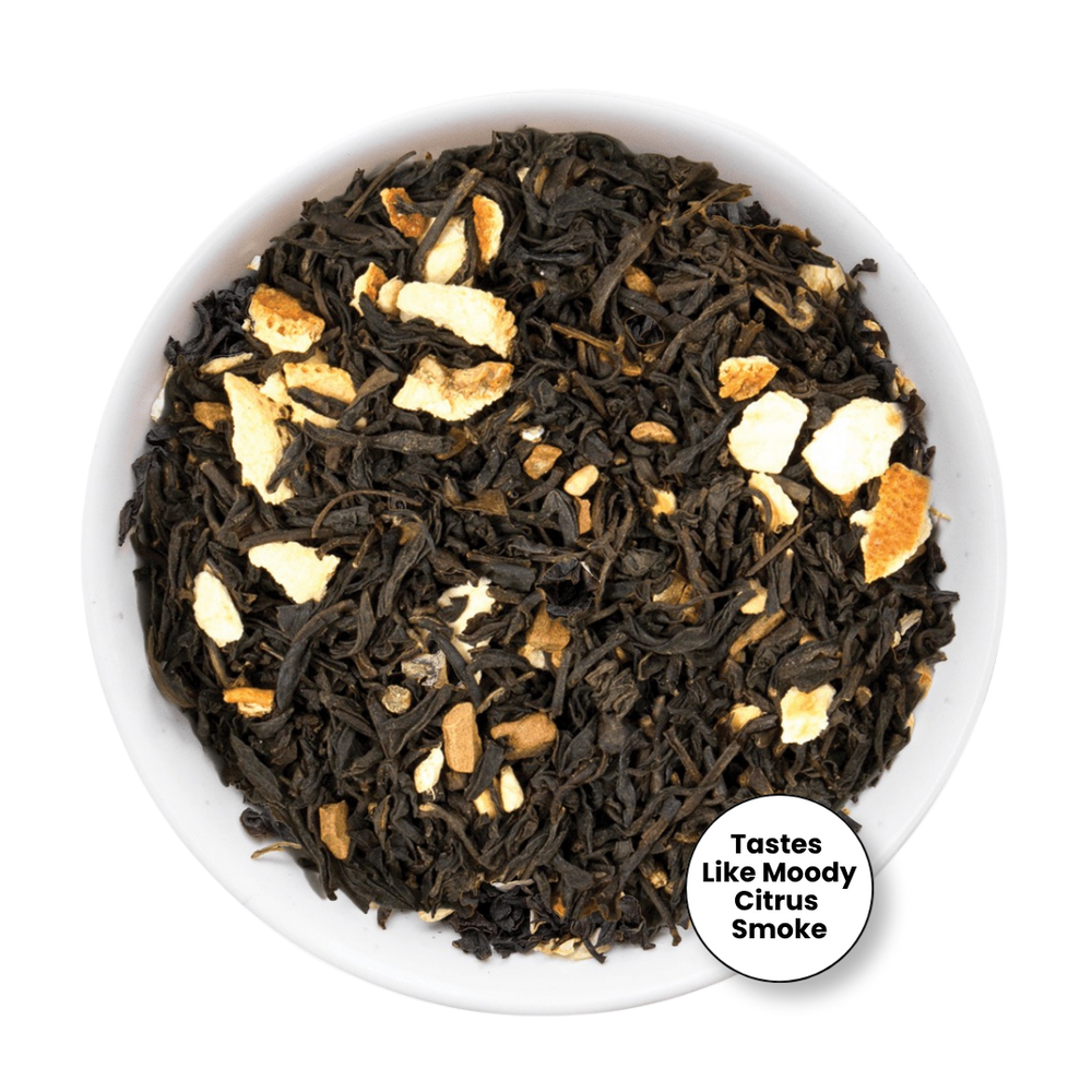 Loose leaf tea blend in a white bowl, featuring dark dried tea leaves mixed with pale orange citrus peel pieces. A white circular badge in the lower right reads 'Tastes Like Moody Citrus Smoke'.