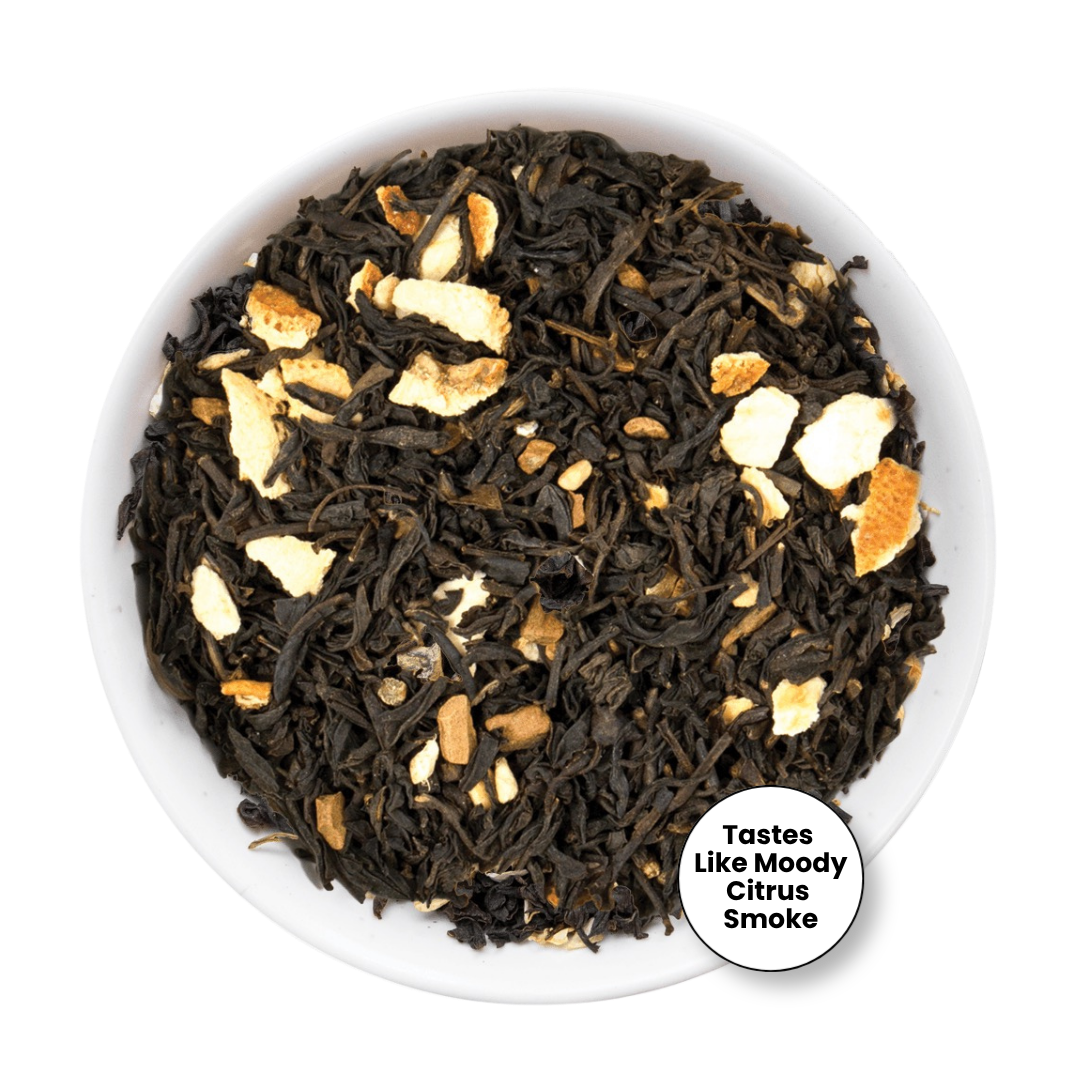 Loose leaf tea blend in a white bowl, featuring dark dried tea leaves mixed with pale orange citrus peel pieces. A white circular badge in the lower right reads &