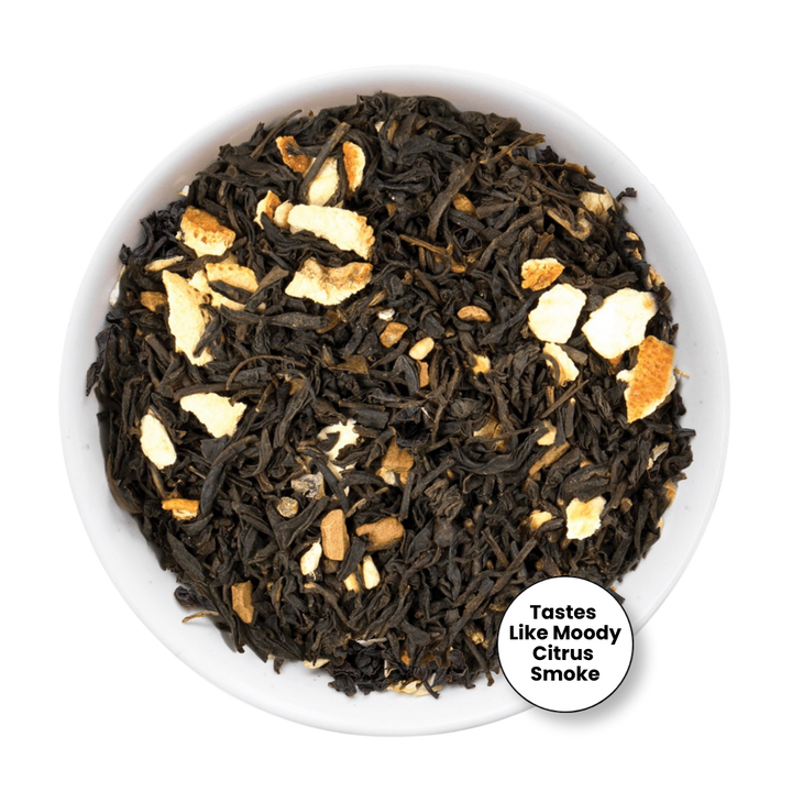 Loose leaf tea blend in a white bowl, featuring dark dried tea leaves mixed with pale orange citrus peel pieces. A white circular badge in the lower right reads &