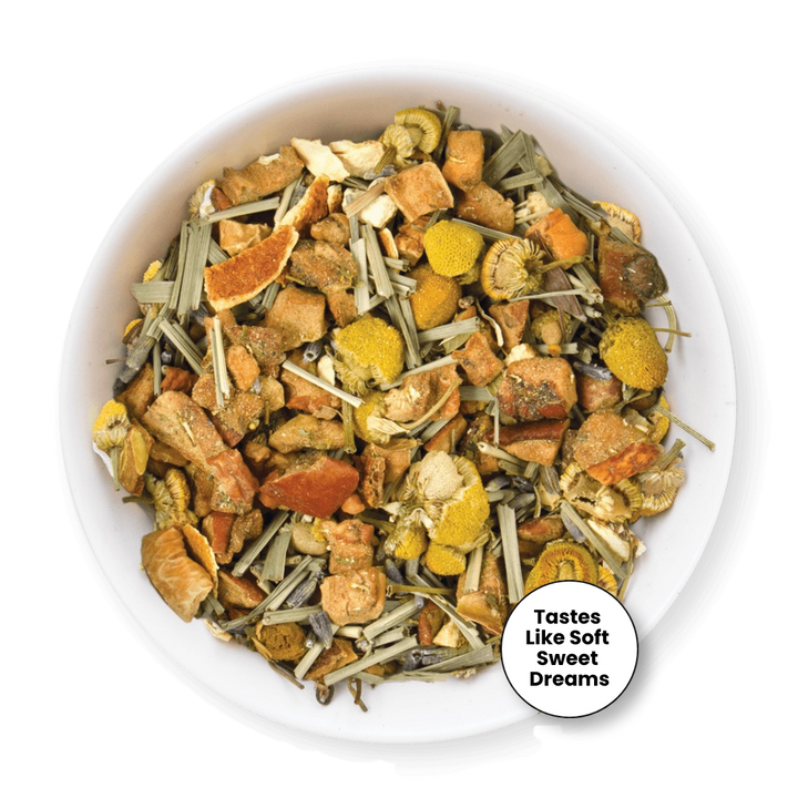 A top-down view of a white bowl filled with a loose herbal tea blend. The tea contains dried orange slices, yellow flower petals, green stems, and brown roots.
